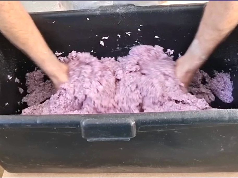 Hände kneten eine lila Masse in einem großen schwarzen Behälter.